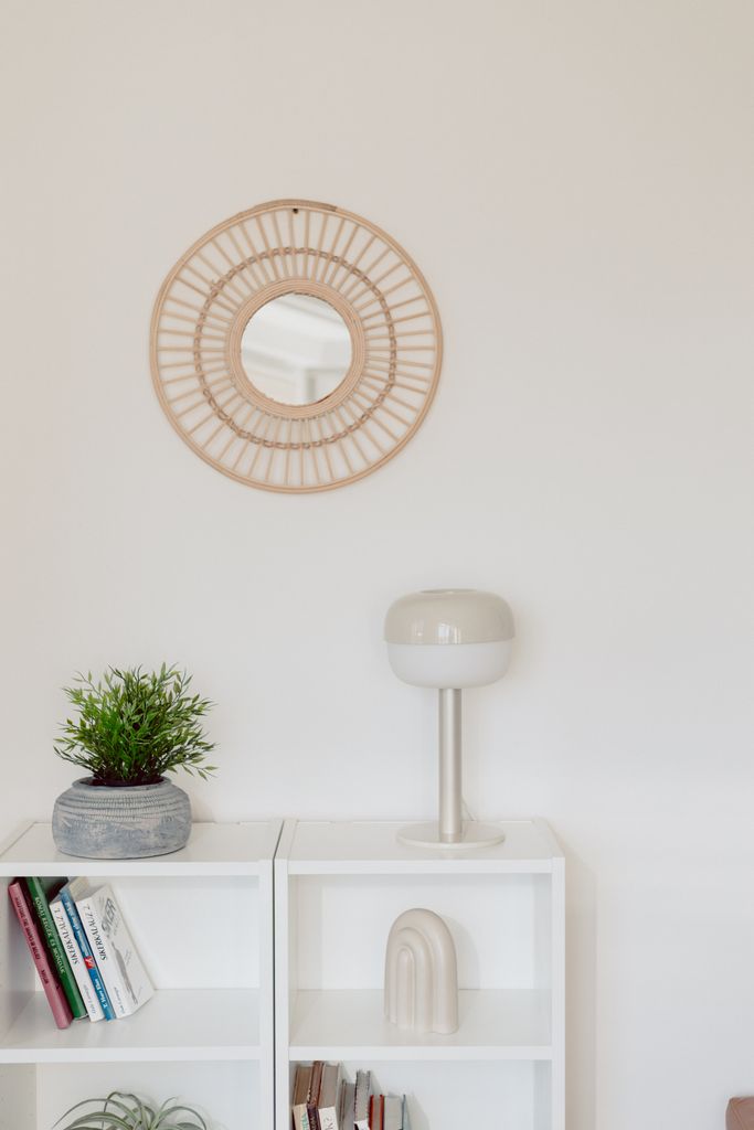 oval woven bamboo wall mirror hung on white wall above white shelving unit with plant and lamp