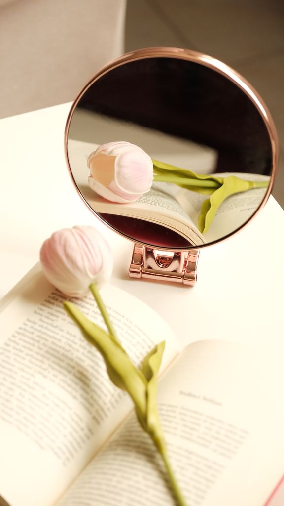 Soft pink tulip reflected in a round mirror on an open book, creating a gentle, serene composition.